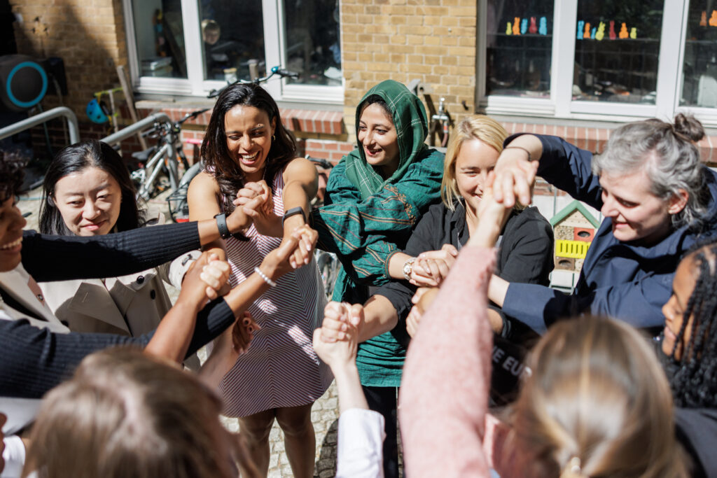 Group of people standing outdoors in a circle, smiling and joining hands together in a team gesture.