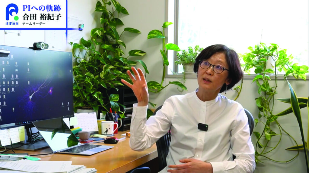 Woman seated at a desk in an office, speaking and gesturing, with a computer and plants in the background and a RIKEN label on screen.