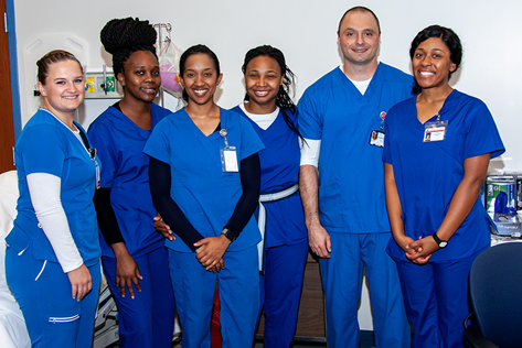 Group of six healthcare workers in blue scrubs standing together in a clinical setting, smiling at the camera.