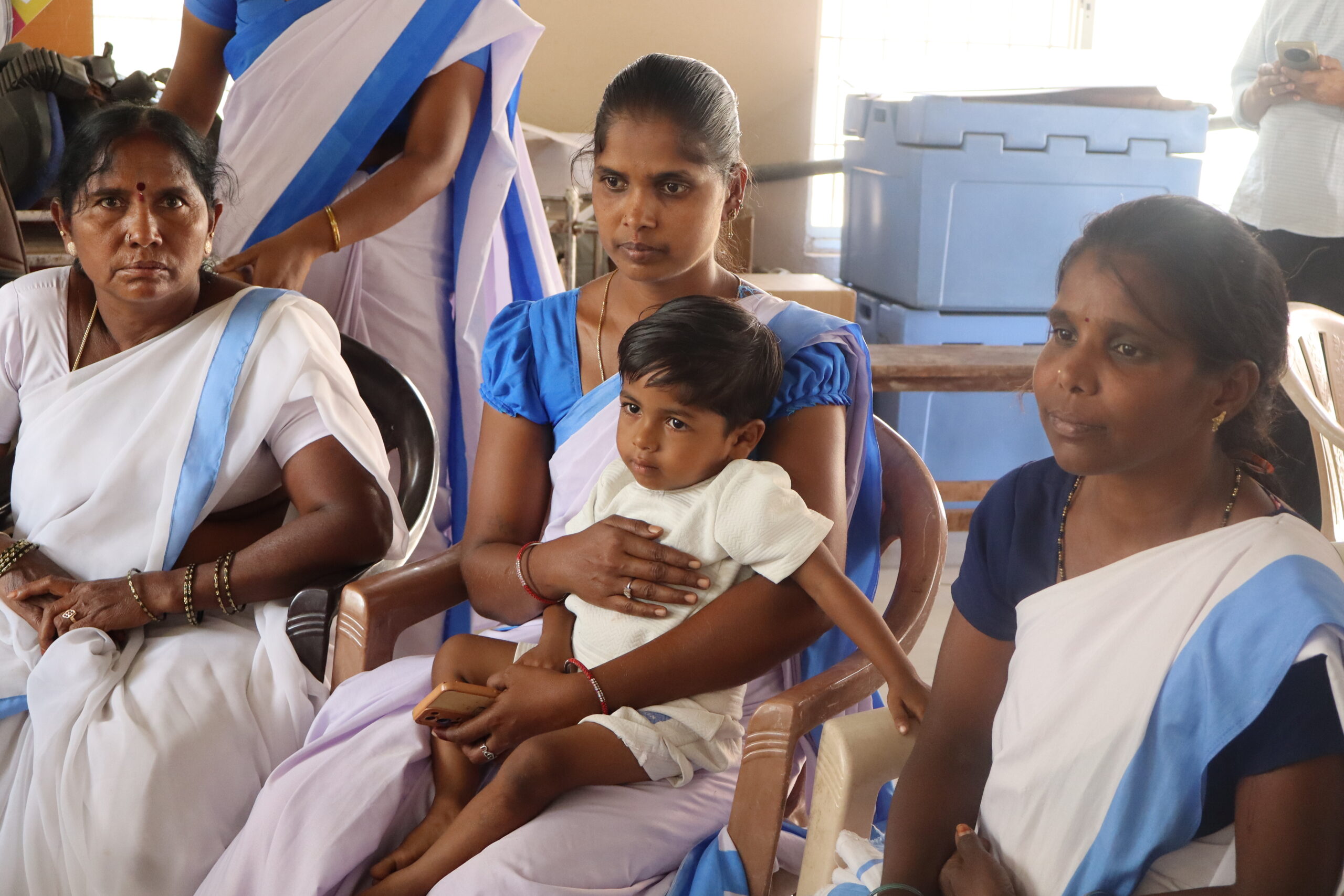 Three women seated indoors during a community session, with one holding a young child, all looking ahead attentively