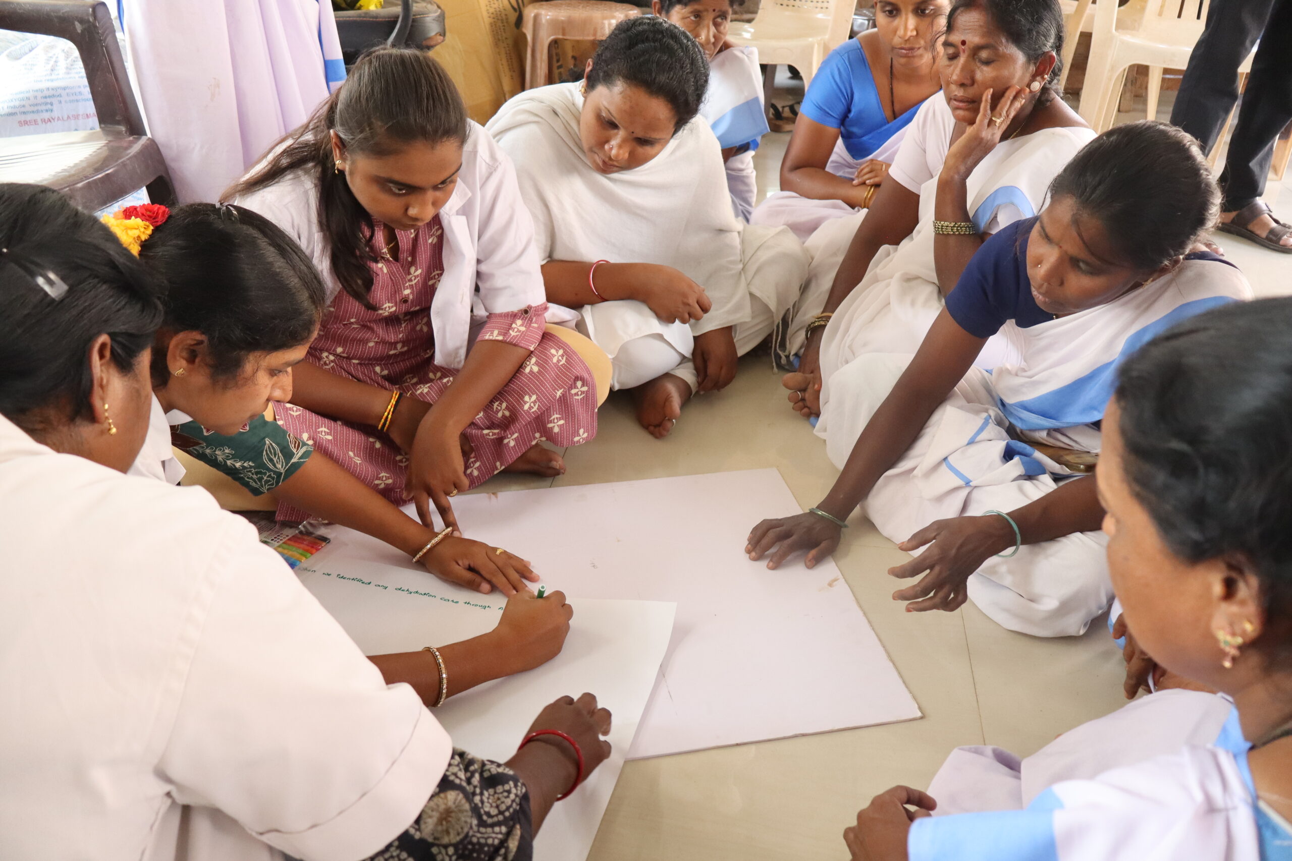 Group of women seated on the floor in a workshop setting, collaborating on a large sheet of paper as one writes and others observe